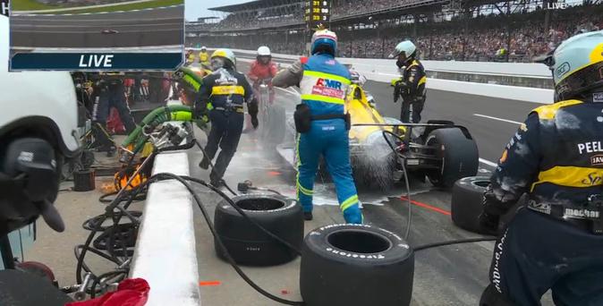 Pit crew and safety team rush to extinguish the fire, flames can still be seen over the pit wall near the fuel pump.