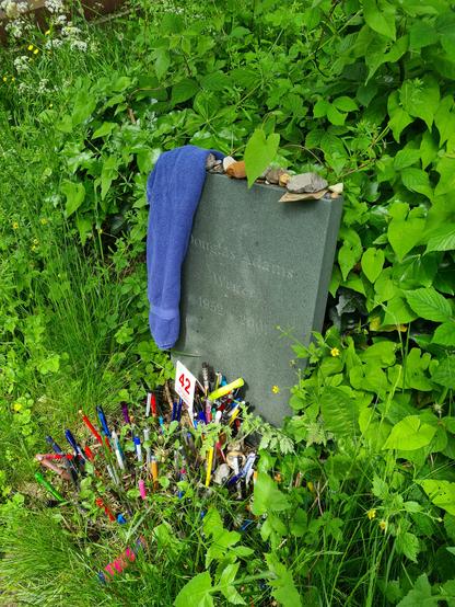 Douglas Adams' grave with blue towel on the gravestone in 2023. Pebbles are stacked on top, there is once again a new box of pens as well as dozens of pens sticking out of the grass in front of that. A small sign with 42 sits in between
