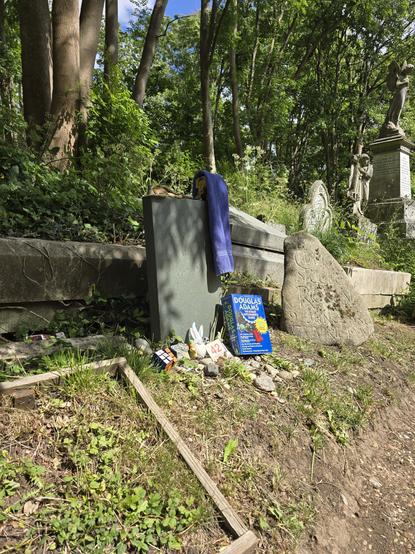 Douglas Adams' grave with blue towel on the gravestone in 2025. The box of pens is there but not visible as my Hitchhiker's book is resting in front of it. The same 42 sign from 2023 is still there. Also pebbles on top with a miniature dalek and a candle holder with this face printed on it. Below is a rubik's cube and a garden gnome.