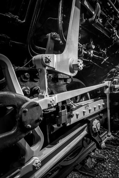 The image is a black and white photograph of a close-up view of a steam locomotive's undercarriage. The focus is on the intricate mechanical components, including a large white metal beam with multiple bolts and rivets, which is part of the locomotive's drive mechanism. The beam is connected to a series of rods and levers, which are essential for transferring power from the engine to the wheels. The wheels are visible in the lower part of the image, with the metal spokes and the flanges that guide the wheels along the tracks. The background shows additional parts of the locomotive, including pipes and other mechanical elements, all set against a dark background that emphasizes the details of the machinery. The photograph captures the complexity and craftsmanship of the steam locomotive's engineering.

Provided by @altbot, generated privately and locally using Ovis2-8B