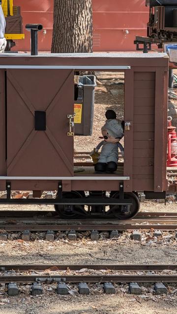 A stuffed Mickey Mouse in an engineer's outfit is sitting inside a tiny boxcar