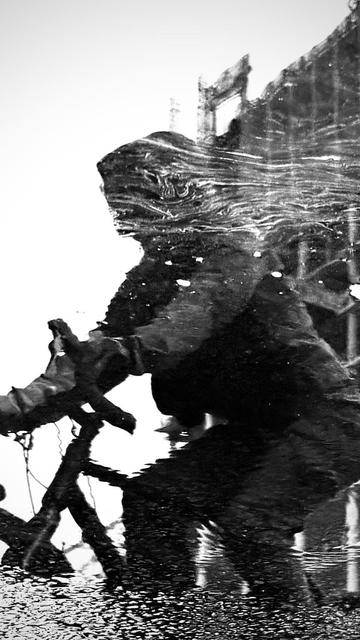 A shadowy reflection of a cyclist and their bike is captured in a puddle, creating an abstract effect. The image is monochromatic, highlighting the ripples of water and the surrounding urban environment.