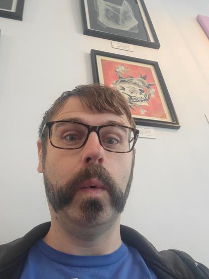 Me, a white guy with a handlebar mustache and a soul patch, making an oooh face. Framed skull art is on the wall behind me in the background.