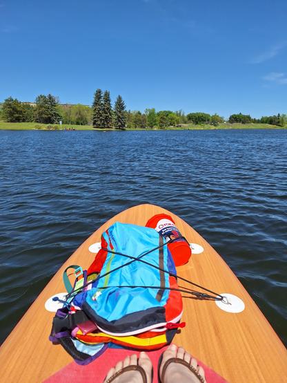 A summery photograph of the front of a wood-coloured stand up paddle board. On it, a colourful backpack and an orange boating safety bucket.

The water is showing small ripples - signs of a good breeze, and in the distance, the grassy shoreline is lined with trees. The sky is clear and blue.