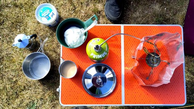 Foto eines Campingtisches von oben aufgenommen. Öinks neben dem Tisch stehen unten ein runder Wassertob, schräg darüber ein Eimer 10%iger Sahnejoghurt und dazwische, ebtwas nach links versetzt eine Espressokanne. Rechts steht ein orangefarbener Campingtisch auf dem oben links eine becherartige Schüssel mit Sahne oder etwas ähnlichem darin steht. Rechts daneben eine Gaskartusche von Primus an die ein Schlauch angeschlossen ist der nach rechts abgeht, sowie eine Tasse mit undurchsichtigem, hellen Inhalt; Ein Topfdeckel liegt ebefalls dabei. Auf der anderen Hälfte des Tisches teht ein Gaskocher der von einer ziemlich deformierten Windschutzvorrichtung aus Blech umgeben ist.