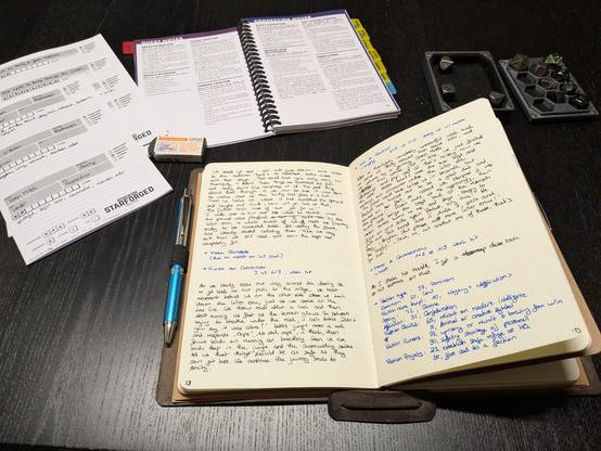 My campaign journal opened on a desk, the current spread completely filled with my hand writing - mostly in black, rolls and such documented in blue. Behind that a bunch of sheets and the reference guide of Starforged, as well as a small dice tray with dice.