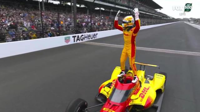 Alex Palou stands atop his race car, arms raised in victory, with the Indianapolis Motor Speedway brick finish line in the background.
