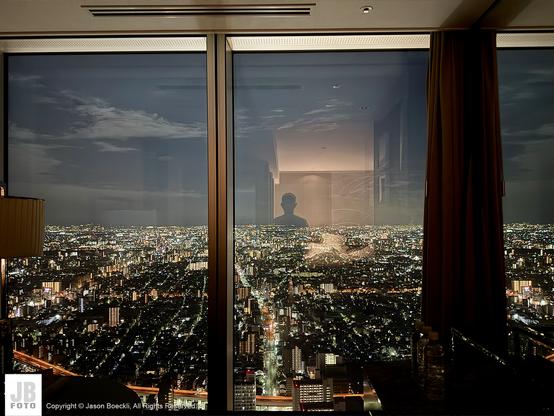 A view of a modern city at night through glass windows.  A person’s reflection is visible on one of the windows.