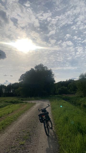 Rower terenowy na szutrowej drodze obok rowu z wodą, słońce na lekko zachmurzonym niebie
