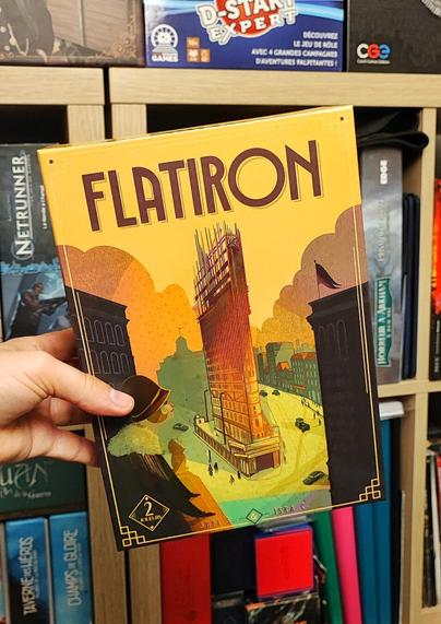 🇨🇵 Boite du jeu de société Flatiron. De forme rectangulaire, aux tons jaunes, verts et noirs. On voit au centre le Flatiron Building en construction à New-York.
En arrière plan de la photo un bout de bibliothèque avec des jeux dedans.
🇬🇧 Flatiron board game box. Rectangular in shape, in shades of yellow, green and black. In the center is the Flatiron Building under construction in New York.
In the background is a bookcase with games inside.