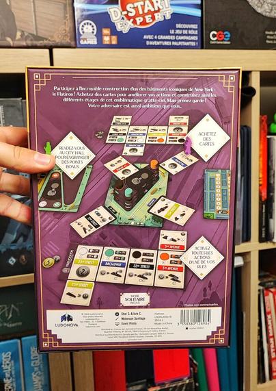 🇨🇵 Dos de la boite du jeu Flatiron. On y voit le matériel et un survol rapide des règles du jeu. Le fond du visuel est de couleur violette.
🇬🇧 Back of the Flatiron game box. It shows the equipment and a quick overview of the game rules. The background is purple.