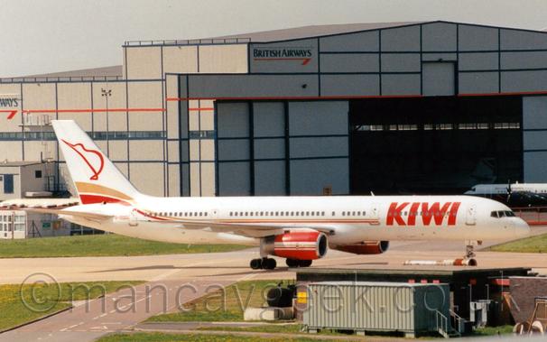 Side view of a white, twin engined jet airliner taxiing from left to right.
The plane has thin gold and red stripes running along the body with large red "Kiwi" titles on the forward fuselage, and a the red outline of a stylised kiwi bird.
The engine pods under the wings are red, with white and gold stripes.
Green grass fills the left of the foreground, with some low buildings and a grey shipping container on the right.
A large grey hangar with blue and red piping and blue British Airways titles dominates the background, with a blue and grey plane parked outside it on the right, and another plane in similar colours inside the open doors.
Grey sky with hints of blue fills the rest of the frame.