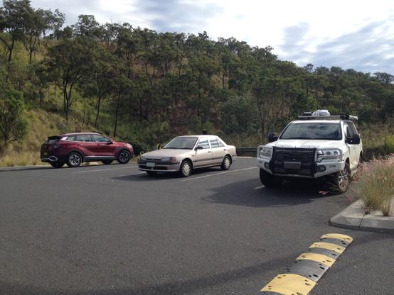 A view of the single width carpark with three cars in foreground, left to right a red Kia Sportage, a silver 1990s Mazda 323 and a white Toyota Landcruiser Prado with offroading kit. Background is the beautiful hillside of Mount Morgan with gum-trees and lime grass bushes scaling the hill.