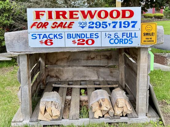 An outdoor firewood stand displaying a sign that reads "FIREWOOD FOR SALE" with pricing details. It lists stacks for $6, bundles for $20, and mentions half and full cords. Neatly stacked firewood is visible beneath the sign