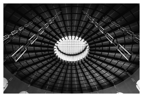 Eine künstlerisch gestaltete Decke dominiert das Bild, die aus konzentrischen, horizontalen und vertikalen Elementen besteht. In der Mitte befindet sich ein großer, runder Ausschnitt, der Tageslicht hereinlässt. Lange, schmale Ketten hängen von der Konstruktion herab und verleihen dem Raum eine gewisse Dynamik. Die monochrome Farbgebung verstärkt die geometrischen Strukturen und schafft eine eindrucksvolle, moderne Atmosphäre.