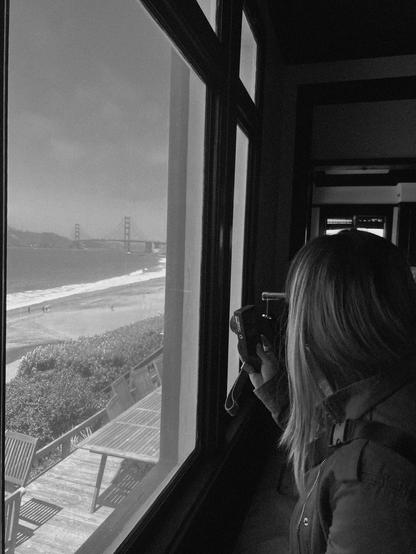 A person stands inside a room, gazing out a large window towards the Golden Gate Bridge in the distance. They hold a camera, capturing the view of the beach and ocean below. The scene is in black and white.