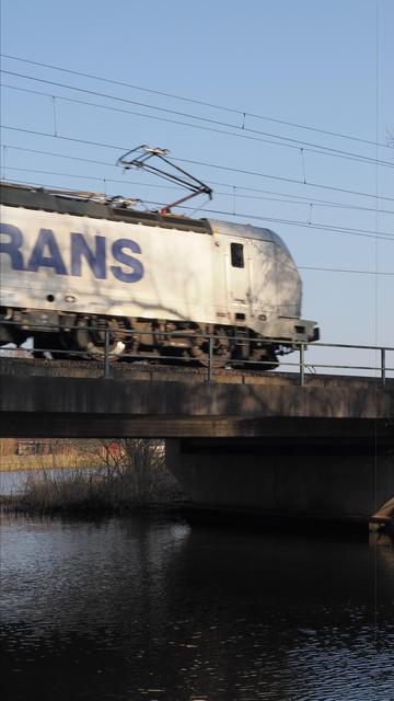 A freight train moves across a bridge over a body of water. The side of the train has large, partially visible lettering. Overhead power lines stretch across the sky, and the water beneath reflects the scene. Grassy and shrub-covered banks line the waterway.