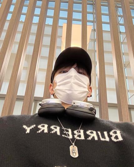 Selfie of Seungmin looking down at the camera, wearing a baseball cap, mask, burberry sweater, and a medallion pendant with a Chrome Hearts dagger on it. He also has over-ear headphones hanging around his neck.