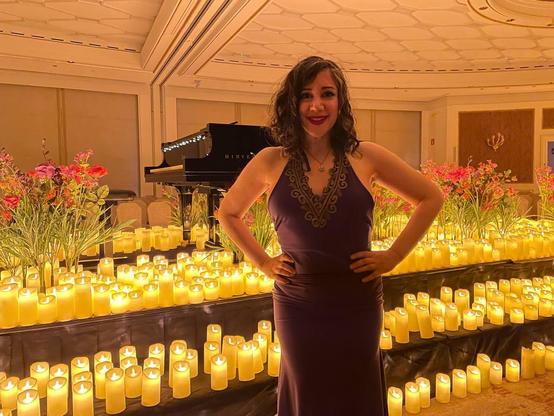 Laura con un vestido de corte sirena morado, con un ribete dorado en el escote en V. Detrás, un piano rodeado de velas y flores.