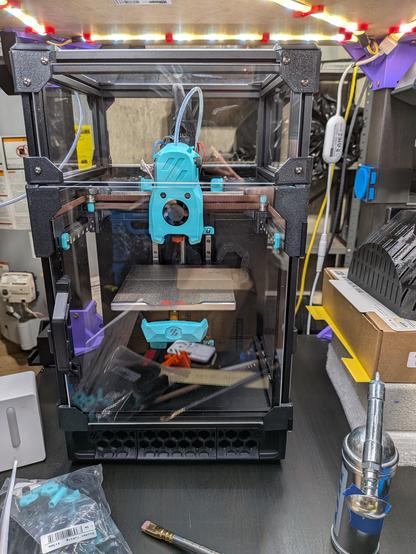 A compact 3D printer sitting on a surface surrounded by assorted tools, boxes, and other materials.