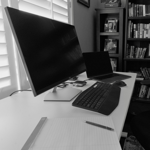 Black and white scene of a laptop and monitor on a white desk in a dimly lit office, with a bookshelf in the background.