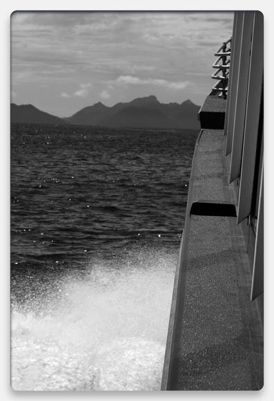 Black and white photo. 
Boat crashing through waves wading towards mountain in the distance.