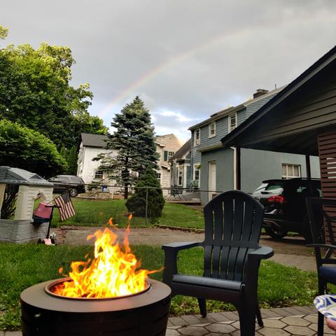 A fire in a "smokeless" fire pit beside an empty, plastic Adirondack chair underneath and overcast sky with a rainbow.