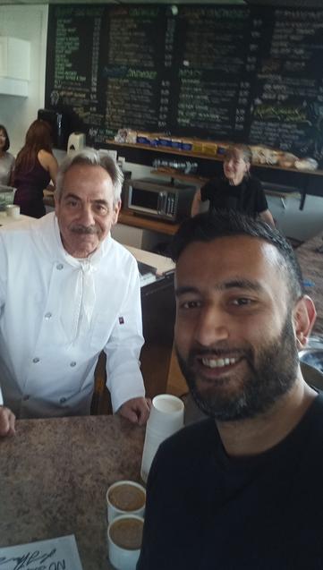Two men posing for a selfie inside a café. The man on the left is wearing a white chef's coat and smiling behind the counter. The man on the right, in a black shirt, is taking the selfie and smiling. Behind them, a woman and another customer are visible, with a large chalkboard menu filling the background. Two cups of coffee sit on the counter in front.
