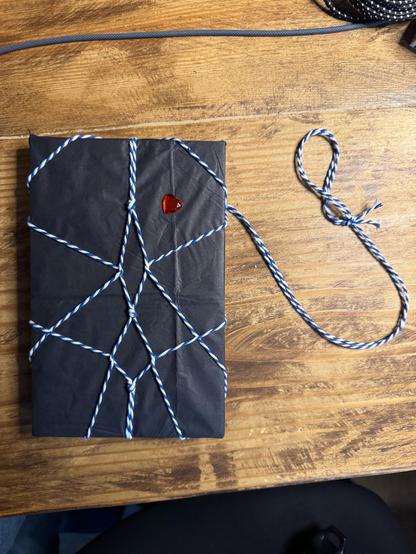 Books from Safeword Press wrapped in black paper, tied shibari-style with white and blue cord. A heart-shaped red gem sits in the upper right section.