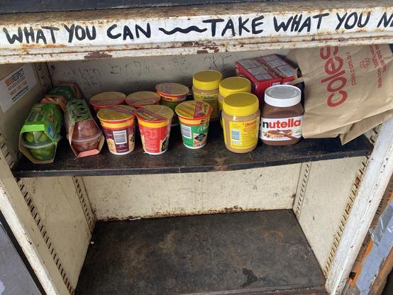 Food cupboard with two shelves; the top one has pantry items like cup noodles, fruit cups, peanut butter and jatz crackers); the bottom shelf is empty. There's writing in black sharpie on the top of the cupboard "[Give] what you can ~ take what you N[eed]."