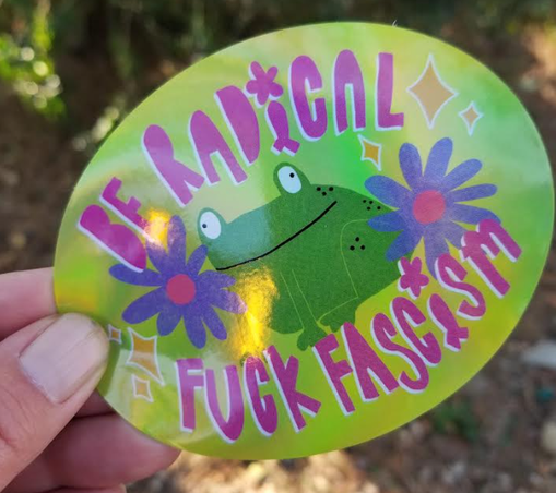 An oval sticker. It features a slightly derpy looking cartoon frog sitting between two purple daisy type flowers. The text is in a hot pink funky font and reads be radical fuck fascism.