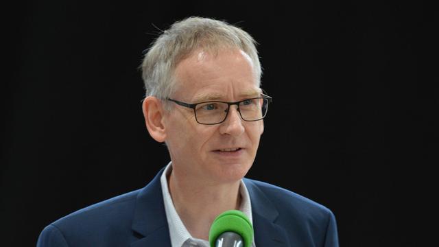 Porträt von Olav Schmidt. Ein Mann mittleren Alters mit kurzen, grauen Haaren und einer Brille. Er trägt einen dunklen Anzug und spricht in ein grünes Mikrofon. Der Hintergrund ist schwarz.