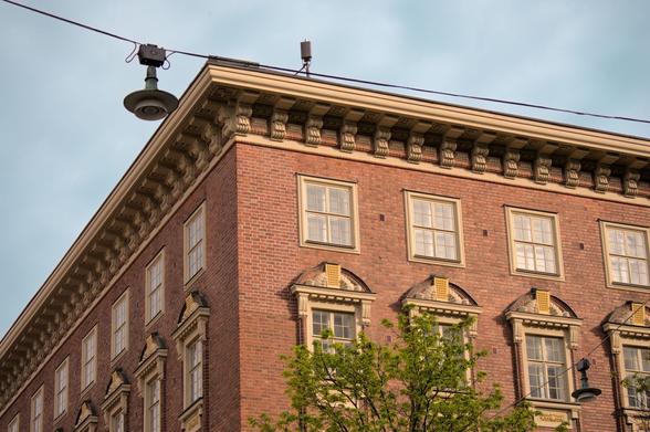 The corner of a brick building lit by the evening sunlight. The building facades has very regularly spaced windows, and is filled with ornaments. In front, cable-suspended street lamps and the top of a tree. On top of the roof is a wireless phone network antenna device.