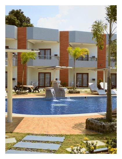 This image depicts a resort with a swimming pool as the central focus. In the foreground, there's a paved area around the pool, leading to some stepping stones and yellow flowers on the right.

The swimming pool is blue and appears to have a small waterfall feature in the middle. Surrounding the pool are lounge chairs, some with umbrellas, and a few small palm trees.

In the background, there is a multi-story building with balconies and a mix of light-colored and orange-red accents on its facade. More lush green trees are visible behind the building and on the left side of the image. - Google Gemini