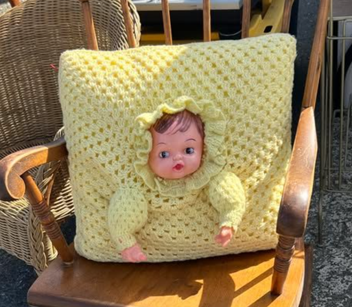 A yellow crochet cushion on a wooden rocking chair.  The crochet is a big granny square. In the middle of the square is a plastic doll face, surrounded by a ruffle like the baby is wearing a bonnet. Under this, two crochet arms protrude from the cushion, each with a plastic doll hand attached.