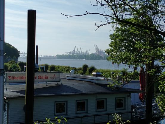 Im Vordergrund ein flaches Gebäude und Sträucher. Dahinter die Elbe, am anderen Ufer Hafenkräne. Auf dem Dach des Gebäudes ein Schild mit roter Schrift: Dübelsbrücker Kajüt.