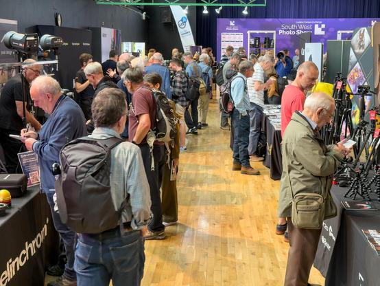 A busy exhibition hall filled with attendees exploring various photography booths and displays, with several people closely inspecting gear and speaking with vendors