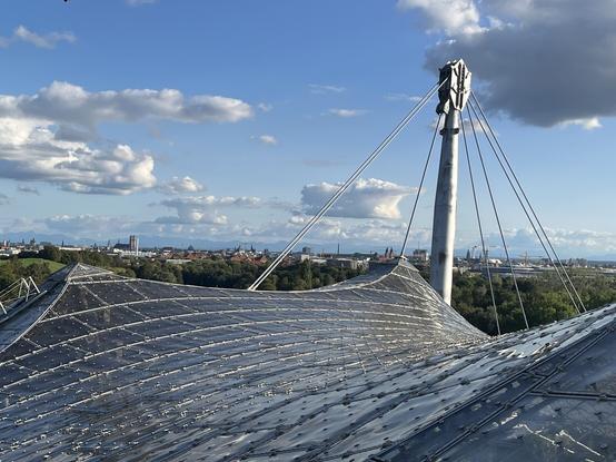 Blick über das Dach des Olympiastadion in München: im Vordergrund mehrere Schwünge und Spitzen des ikonischen Glasdachs, dahinter ein Tragepfeiler, von dem aus mehrere Stahlseile das Dach halten. Im Hintergrund ein Park und Stadtsilhouette, darüber blauer Himmel mit einzelnen Wolken.