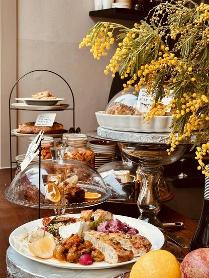 Buffet spread featuring multiple platters of food, including a large white plate with a variety of appetizers, smaller plates on a tiered stand, and a display of baked goods under glass domes, with yellow mimosa flowers decorating the scene.