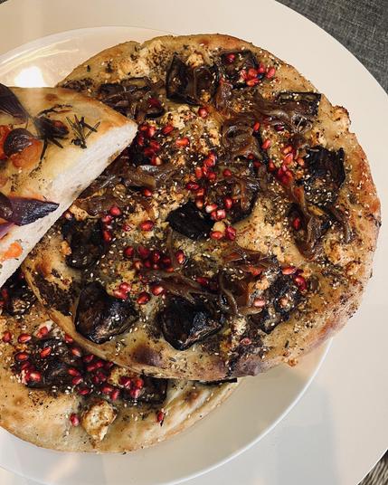 Two focaccia breads on a white plate, one topped with caramelized onions, dark roasted vegetables, feta, sesame seeds, and pomegranate seeds, with a slice of another focaccia with herbs and red onion visible.