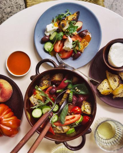 A table set with two plates of panzanella salad featuring bread chunks, tomatoes, cucumbers, olives, and cheese, with a bowl of red sauce, bread sticks, and a peach nearby.