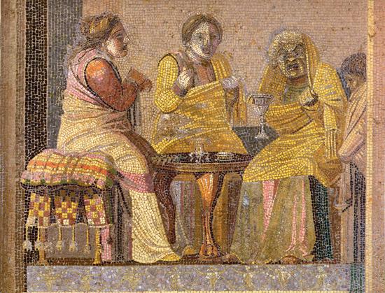 Mosaic panel depicting a theatre scene of two women making a call on a witch, each actor wears a theatre masks. The women are shown in yellow-gold gowns sitting on cushioned chairs while a fourth figure is partially obscured by a pillar or the border of the frame. The actors sit around a small table and the witch holds a cup. The piece is credited to Dioscorides of Samos.