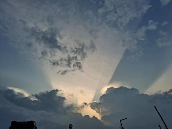 A dramatic sky during sunset with dense clouds partially covering the sun, casting long rays of light through the atmosphere. The scene includes silhouetted objects like poles and a rooftop in the foreground, adding depth to the moody and serene sky.