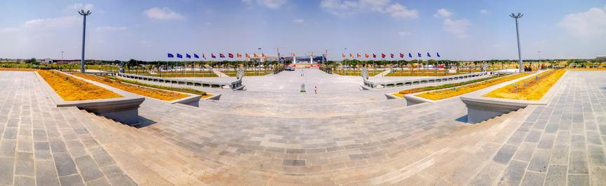 Photo shows a wide panorama from the platform where the main stele is placed, towards the main road. On the other side of the road, there is a large arena being built. The platform has three main levels, with wide stairs to walk up between them. On the side of the stairs there are wide flowerbeds, a bit dry in this early photo. Two tall lampposts with floodlights meant for nighttime decorative lights of the monument. On the ground square there are a two guardian Naga-statues in classic design where their tails are rails framing in the main platform. Two statues are visible on the main square, together with one person on the other site of the square there are more flower beds, and two sets of flagpoles with the national flag and the Buddhist flag. The background is a light blue sky with a few scattered clouds.