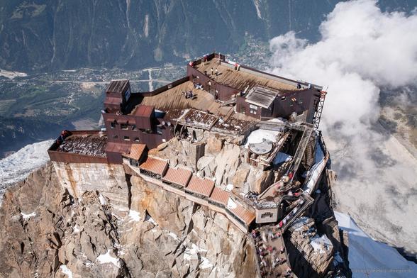 Die Bergstation an der Aiguille du Midi, fotografiert von der Gipfelhöhe von 3842 m. Die Gebäude der Bergstation schmiegen sich an einen Felsblock des Vorgipfels. Eine Gruppe von Touristen nähert sich auf einem Steg, der außerhalb des Bildfeldes verläuft, während einige Personen eine Treppe hinaufgehen. Durch die längere Belichtungszeit erscheinen die Konturen der entfernten Menschen verwischt, was dem Bild eine dynamische Wirkung verleiht und den Eindruck erweckt, dass die Gruppe hastig die Treppe hinaufsteigt. Auf dem Bergstationskomplex ist eine Aussichtsplattform sichtbar, auf der einzelne Personen stehen. Der gesamte Felskomplex mit der Bergstation ragt von unten empor, sodass in der Ferne die Stadt Chamonix, etwa 2000 Meter tiefer, erkennbar ist. Rechts im Bild ist eine tiefere Wolkenschicht zu sehen.
