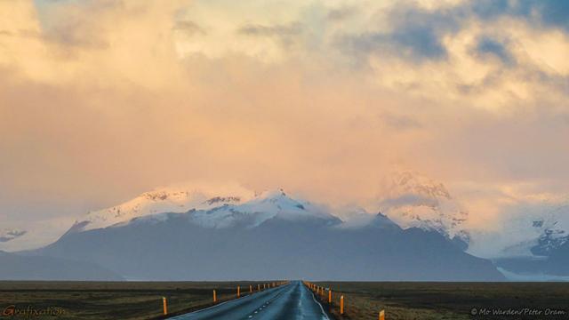 A colour photo of a mountain range at sunset. The mountains are all snow-capped and dropping between them to ground level are glaciers. The foreground is a tarmac road with snow poles and brown vegetation on either side. The cloudbank above the peaks is tinted orange and yellow by the setting sun, and there are cloud shadows across the flanks.
