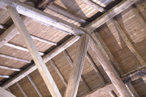 Das Bild zeigt das Innere eines Gebäudes, wobei der Schwerpunkt auf der Decke und den tragenden Balken liegt. Die Decke besteht aus horizontal angeordneten Holzbrettern, die eine natürliche, hellbraune Farbe und eine sichtbare Maserung aufweisen. Die Balken sind dick und robust und weisen eine raue Textur auf, was darauf hindeutet, dass sie aus großen Stämmen gefertigt sind. Sie kreuzen sich in verschiedenen Winkeln und bilden ein komplexes Netz von Stützen. Die Balken sind hauptsächlich vertikal und horizontal angeordnet, wobei diagonale Verstrebungen der Struktur Stabilität verleihen. Die Beleuchtung des Bildes ist natürlich und wirft Schatten, die die Textur und Tiefe des Holzes hervorheben.