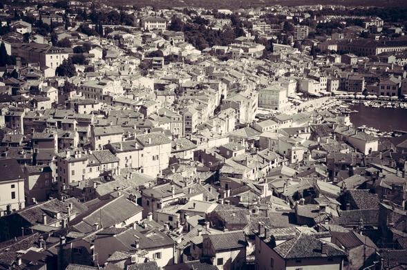 Das Bild zeigt ein dicht bebautes Stadtgebiet, wahrscheinlich eine historische Stadt, aus einer erhöhten Perspektive. Der Sepia-Ton verleiht dem Foto einen Hauch von Vintage. Die Gebäude stehen dicht beieinander und weisen verschiedene Dachformen auf, überwiegend mit Ziegeln und einige mit Schornsteinen. Die Architektur deutet auf einen europäischen Einfluss hin, mit Gebäuden unterschiedlicher Höhe und Gestaltung, einige davon mit verschnörkelten Fassaden. Im Hintergrund ist ein Gewässer mit mehreren angedockten Booten zu sehen, was auf einen Hafen oder Jachthafen hindeutet. Der Grundriss der Stadt ist unregelmäßig, mit schmalen Straßen, die sich zwischen den Gebäuden schlängeln. Der Gesamteindruck ist der einer geschäftigen, altmodischen Stadt mit einem reichen architektonischen Erbe.