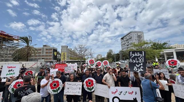 A large crowd is gathered for a protest featuring various signs and banners with messages addressing issues related to Gaza and human rights. Some signs display red flowers, and the scene is set against a backdrop of buildings and a partly cloudy sky.
