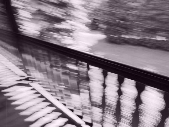 Blurred motion effect capturing a floor with ornate railings on the left, transitioning into a grassy outdoor area. Sunlight creates patterns on the floor, while trees are visible in the background. Image has a monochromatic, sepia like tone.